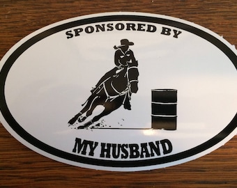 May include: White oval sticker with black text and a silhouette of a person riding a horse around a barrel. The text reads "Sponsored by My Husband".