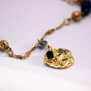 Puede incluir: Un collar de oro con un colgante grande de forma irregular. El colgante est&aacute; decorado con peque&ntilde;as piedras negras y marrones. El collar tambi&eacute;n tiene cuentas azules y doradas.