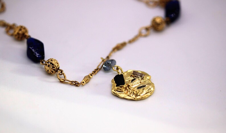 Puede incluir: Un collar de oro con un colgante grande de forma irregular. El colgante est&aacute; decorado con peque&ntilde;as piedras negras y marrones. El collar tambi&eacute;n tiene cuentas azules y doradas.