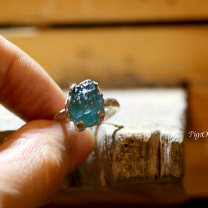 Puede incluir: Un anillo de plata con una gran piedra preciosa azul verdosa sin pulir. El anillo está colocado sobre una superficie de madera.