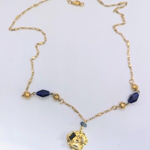 Puede incluir: Un collar de cadena dorada con un colgante dorado y cuentas de vidrio azul. El colgante tiene una forma irregular y texturizada.