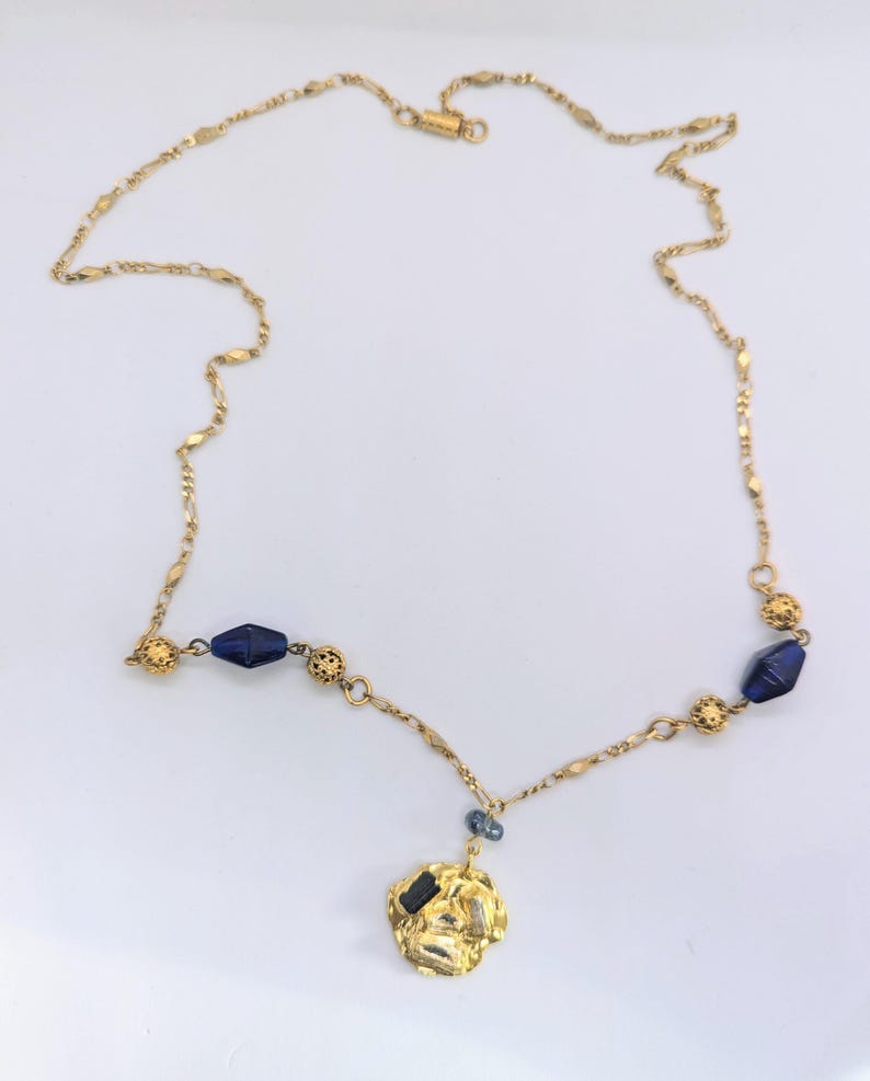 Puede incluir: Un collar de cadena dorada con un colgante dorado y cuentas de vidrio azul. El colgante tiene una forma irregular y texturizada.