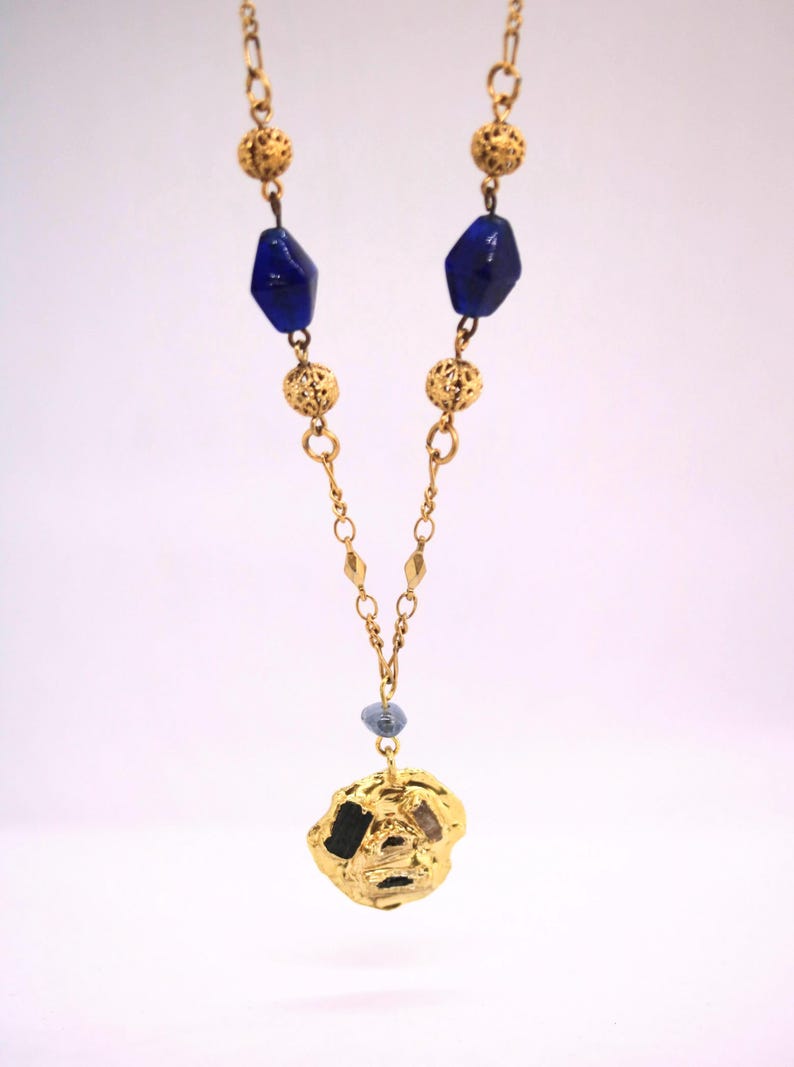 Puede incluir: Un collar de oro con un gran colgante de oro texturizado. El collar est&aacute; hecho de una cadena con peque&ntilde;as bolas de oro y cuentas de vidrio azul.