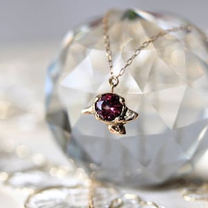 Pode incluir: Um colar com pingente dourado com uma pedra preciosa vermelha escura. O pingente tem uma configuração ornamentada com pequenas pedras de destaque. O colar é exibido em uma esfera de cristal facetada, com um fundo suave e desfocado.