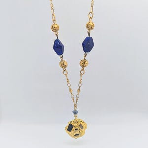 Puede incluir: Un collar de cadena dorada con cuentas de vidrio azul y un colgante dorado. El colgante tiene una forma irregular y texturizada.