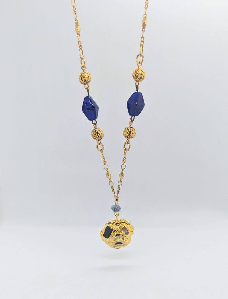 Puede incluir: Un collar de cadena dorada con cuentas de vidrio azul y un colgante dorado. El colgante tiene una forma irregular y texturizada.