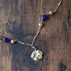 Puede incluir: Un collar de cadena dorada con un colgante de vidrio azul y cuentas doradas. El colgante tiene una forma irregular y texturizada.