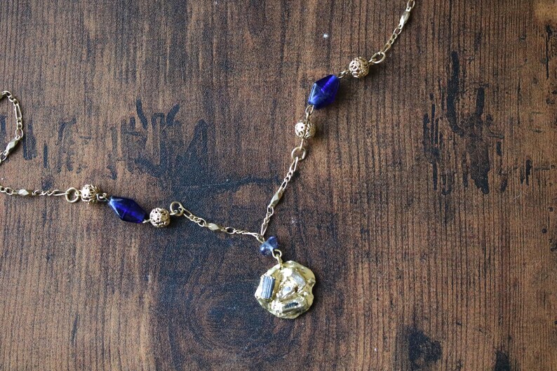 Puede incluir: Un collar de cadena dorada con un colgante de vidrio azul y cuentas doradas. El colgante tiene una forma irregular y texturizada.