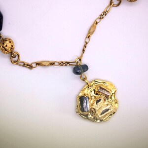 Puede incluir: Collar de cadena dorada con una cuenta de vidrio azul, una cuenta dorada y un colgante dorado con superficie texturizada y piedras negras.