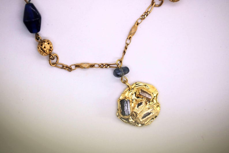 Puede incluir: Collar de cadena dorada con una cuenta de vidrio azul, una cuenta dorada y un colgante dorado con superficie texturizada y piedras negras.