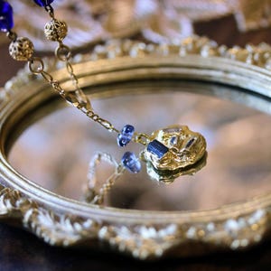 Puede incluir: Un collar de oro con un colgante azul y dorado cuelga de un espejo ovalado dorado adornado. El colgante tiene una forma irregular texturizada con piedras azules.