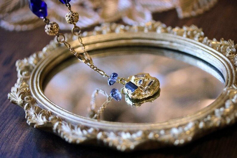 Puede incluir: Un collar de oro con un colgante azul y dorado cuelga de un espejo ovalado dorado adornado. El colgante tiene una forma irregular texturizada con piedras azules.