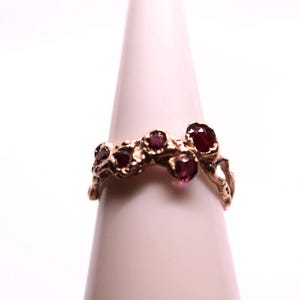 Pode incluir: Um anel de ouro com um grupo de pedras preciosas vermelhas engastadas em uma banda texturizada.