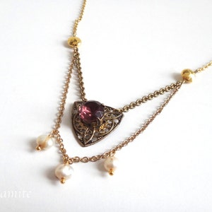 Puede incluir: Un collar de cadena dorada con un colgante triangular que presenta una piedra preciosa púrpura y detalles filigranados intrincados. El collar tiene tres gotas de perlas que cuelgan de la parte inferior.