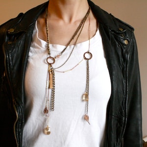 Puede incluir: Un collar de capas con múltiples cadenas y dijes, incluyendo un dije largo con un cristal en el extremo. El collar se lleva sobre una camiseta blanca y una chaqueta de cuero negra.