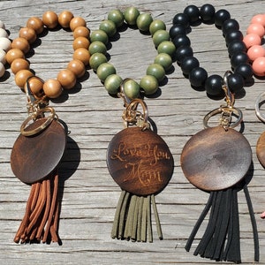 May include: Five wooden beaded bracelets with a round wooden charm and a tassel. The bracelets are in white, brown, green, black, and pink. The charms are engraved with "Love You Mom" on one of them.