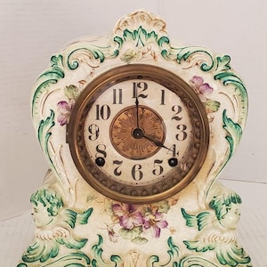 May include: Antique porcelain mantle clock with a white and green floral design. The clock face has Roman numerals and a gold-toned frame. Two cherubs are on the sides of the clock.