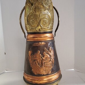 May include: A copper and brass boot scraper with a decorative top and a scene of people sitting at a table. The scraper has a handle on each side.