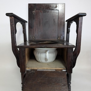 May include: Antique wooden commode chair with a dark finish. The chair features a rectangular back, armrests, and a seat with a circular opening. A white ceramic chamber pot is visible inside the chair's compartment. The chair has a small, hinged door at the bottom.