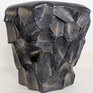 May include: A stool with a cylindrical shape featuring a dark, glossy finish. The exterior is composed of numerous overlapping, irregular, shell-like segments in shades of charcoal, black, and muted bronze, creating a textured, faceted appearance.