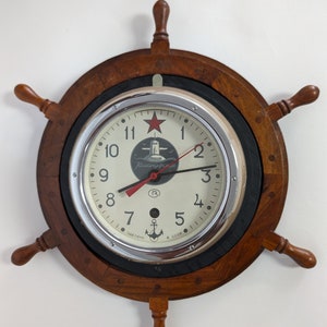 May include: A wooden ship's wheel clock with a white face and black hands. The clock face has a red star and the text "Командирские" in Cyrillic script. The clock is made of wood and has a black background.