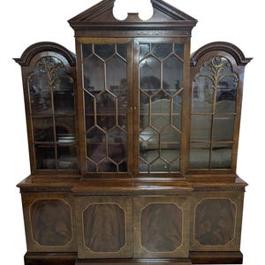 May include: A dark wood cabinet with glass-paned doors and decorative trim. The cabinet has a central section with a triangular top and two side sections with arched tops. The lower section has paneled doors with gold trim.