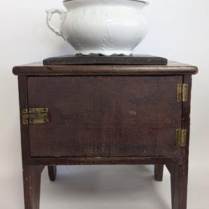 May include: A white porcelain chamber pot with a lid, resting on a dark wooden stand with a small cabinet and brass hardware. The chamber pot has a floral design and handles. The stand has four legs and a single door.