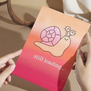 May include: A greeting card with a gradient background of pink and orange, featuring a cartoon snail illustration. The text "still loading..." is printed on the card. The card is held by two hands.