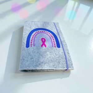 Könnte beinhalten: Graue Filz-Notizbuchhülle mit einem blau-rosa Regenbogen-Design und einem rosa Band. Sie hat einen lila Gummibandverschluss. Das Notizbuch liegt auf einer weißen Oberfläche.
