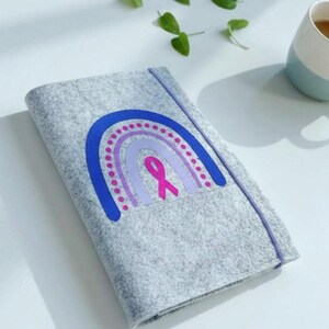 May include: A gray felt notebook cover with a rainbow design in blue, purple, and pink, and a pink ribbon. The cover has a purple elastic band closure.