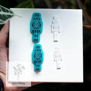 May include: Two blue and two white plastic nutcracker cookie cutters on a white surface. The blue cutters are in the shape of a nutcracker soldier. The white cutters are smaller versions of the same design. The image includes the text "PETALS & PROVISIONS".