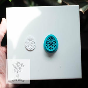 May include: A white, embossed egg-shaped stamp and a teal egg-shaped stamp with a floral design. The teal stamp is next to the embossed stamp. A small square with the text "Petals & Provisions" and a floral illustration is in the lower left corner.