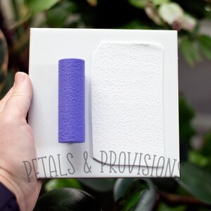 May include: A white textured sheet of paper with a purple textured roller on a white background. The text "Petals & Provisions" is printed on the bottom of the sheet.