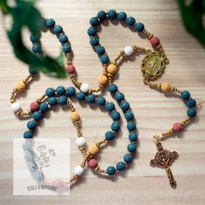 May include: A rosary with blue, white, yellow, and pink lava stone beads. The rosary features a gold-colored crucifix and a decorative centerpiece. The text "Petals & Provisions" is visible on a small card.