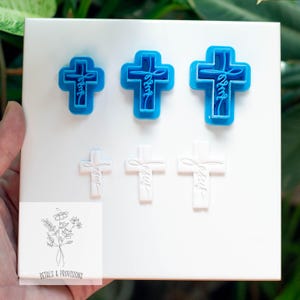 May include: A set of six cross-shaped cookie cutters in two sizes. Three are bright blue, and three are white. Each cross has the word "Jesus" inscribed. The cutters are on a white surface.