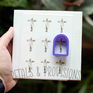 May include: A white sheet of paper with nine images of a cross with a figure on it. The images are arranged in a 3x3 grid. A purple plastic cookie cutter in the shape of a cross is on top of the sheet of paper. The text "PETALS & PROVISIONS" is at the bottom of the sheet of paper.