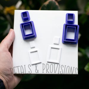May include: A white tile with several blue and white rectangular cookie cutters. The blue cutters are nested, and the white cutters are single pieces. The text "PETALS & PROVISIONS" is printed in gray at the bottom of the tile.