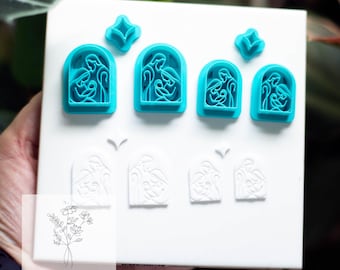 May include: A white rectangular display board holds several teal and white polymer clay cutters. The teal cutters are in the shape of arched windows with a nativity scene inside. The white cutters are also arched with a similar design. The board has the text "Petals & Provisions".