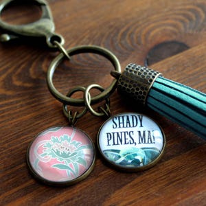 May include: A keychain with bronze-coloured clasp, rings, and charms. One charm has a floral design, and the other says "Shady Pines, MA!". A teal tassel hangs from the keychain. The background is a wooden surface.