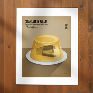 May include: A framed art print depicting a black stapler inside yellow jelly on a white plate. The text "STAPLER IN JELLO" and "The Office" are above the image, with the year "2005" on the right.