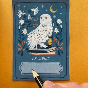 Blue bookplate featuring a snowy white owl sitting on a stack of books with an oil lamp, surrounded by white flowers, a moon and orange berries.
