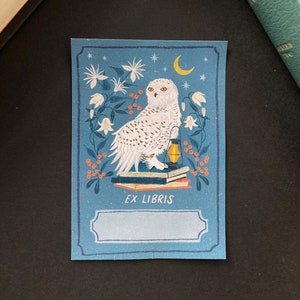 Blue bookplate featuring a snowy white owl sitting on a stack of books with an oil lamp, surrounded by white flowers, a moon and orange berries.