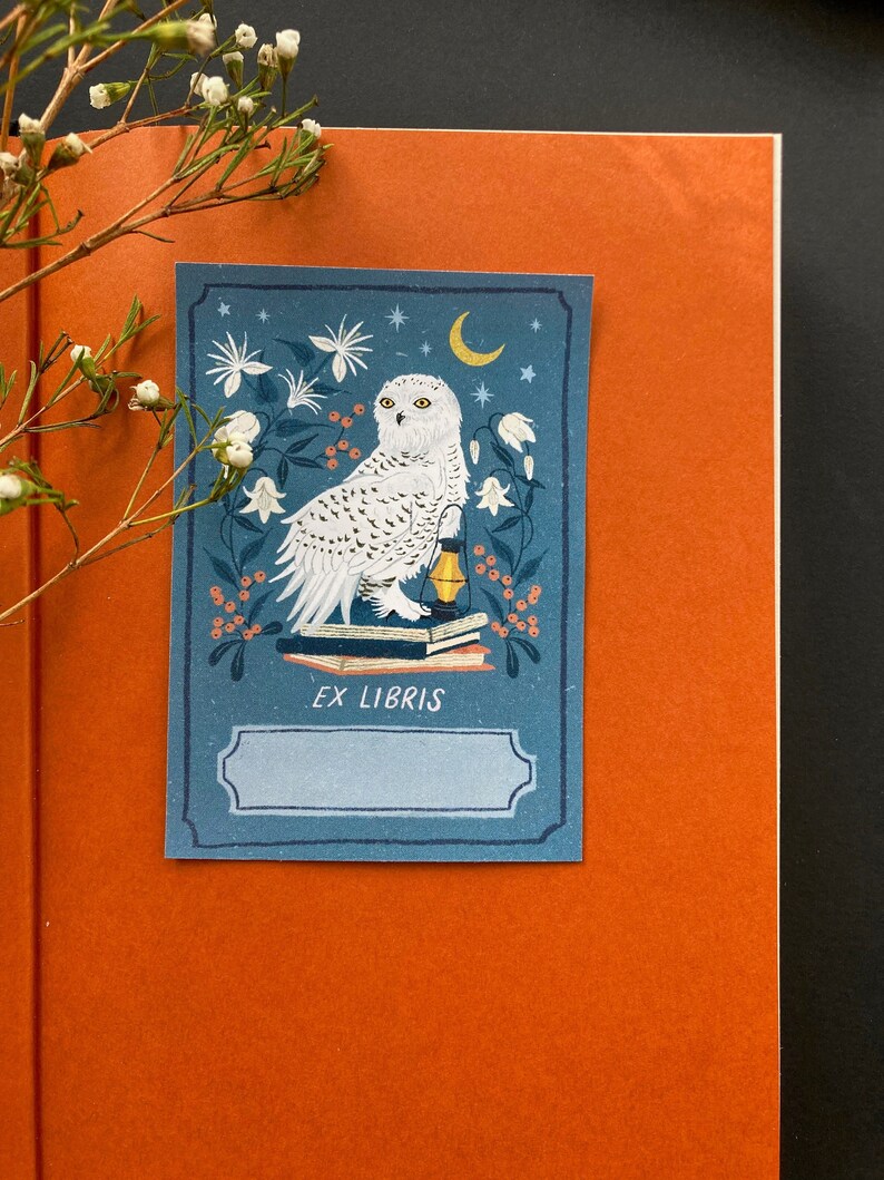 Blue bookplate featuring a snowy white owl sitting on a stack of books with an oil lamp, surrounded by white flowers, a moon and orange berries.