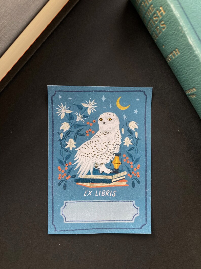 Blue bookplate featuring a snowy white owl sitting on a stack of books with an oil lamp, surrounded by white flowers, a moon and orange berries.