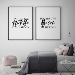 May include: Two black framed prints with white backgrounds and black text. The text on the left print reads "You are the Nutella to my spoon". The text on the right print reads "You are the bacon to my eggs". The prints are hanging on a white wall above a gray bed with pink pillows.