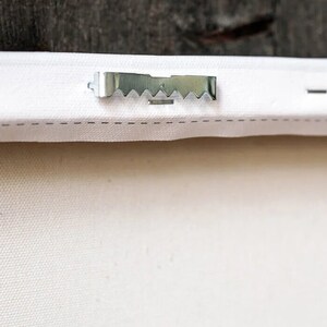 May include: A close-up of a silver metal sawtooth picture hanging hook attached to the back of a white canvas.