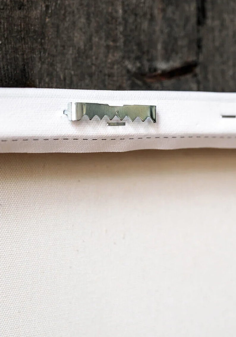 May include: A close-up of a silver metal sawtooth picture hanging hook attached to the back of a white canvas.