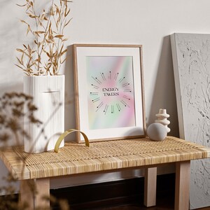May include: A framed print with a rainbow background and black text that reads "Energy Takers". The print is on a wooden table with a woven top and a white vase with dried branches.
