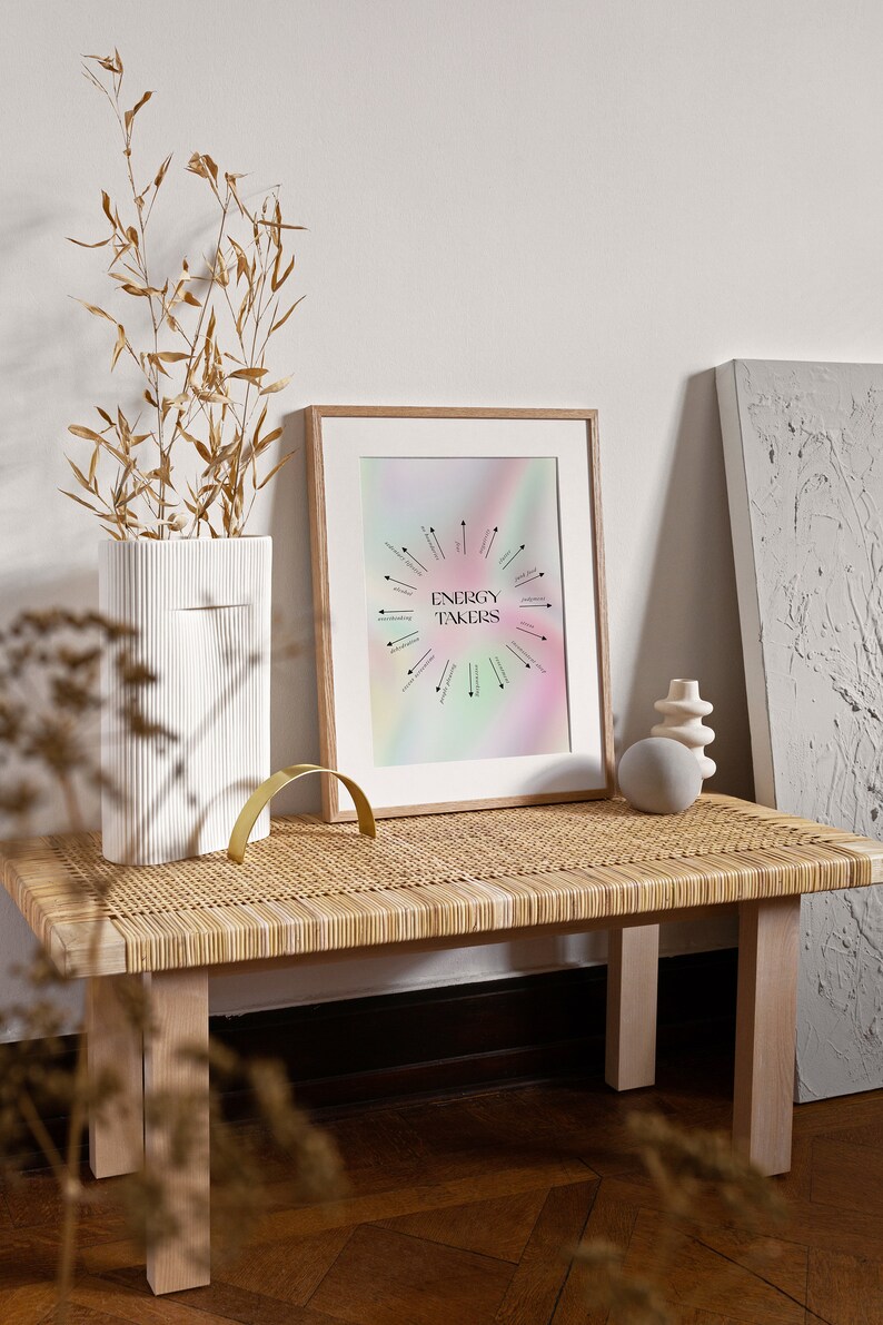 May include: A framed print with a rainbow background and black text that reads "Energy Takers". The print is on a wooden table with a woven top and a white vase with dried branches.