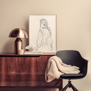 K&ouml;nnte beinhalten: Ein schwarzer Stuhl mit einer beigefarbenen Strickdecke dar&uuml;ber steht vor einer Kommode aus Holz. Eine goldene Lampe mit einem wei&szlig;en Schirm steht auf der Kommode neben einer gerahmten Schwarzwei&szlig;-Zeichnung einer Frau.
